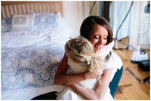 Bride hugging flower girl dressed in white sitting at the edge of a bed.