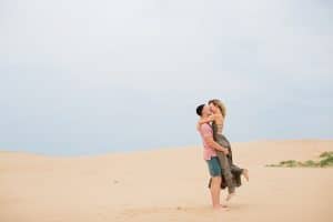 Couple is kissing under the blue sky at Silver Lake Sand Dunes. Soon to be Bride is lifted up by her Fiance in a loving manner.