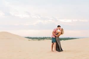 Couple kissing at Silver Lake Sand Dunes for their Engagement Photos. Couple is Standing in the middle of a Dune with the blue sky and some green trees in the background.