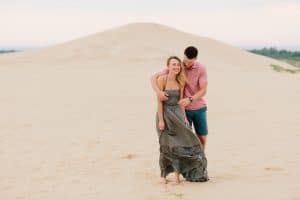 Couple pauses on the dunes at Silver Lake. Soon to be groom is holding his bride from behind while she hunches over and laughs. Couple is posing for their engagement photos.