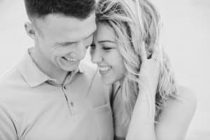 Black and white photo of engaged couple at Silver Lake Sand Dunes. Couple is looking down and smiling while standing close to each other.