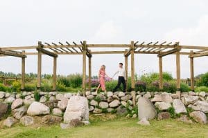 Cherry Point Farms engagement photography session in the summer where couple is walking and holding hands under a wooden arch with picturesque stones