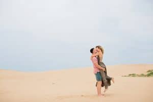 Couple is kissing under the blue sky at Silver Lake Sand Dunes. Soon to be Bride is lifted up by her Fiance in a loving manner.