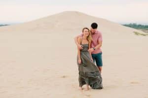 Couple pauses on the dunes at Silver Lake. Soon to be groom is holding his bride from behind while she hunches over and laughs. Couple is posing for their engagement photos.