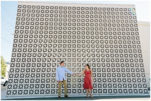 Couple standing in front of an intricate wall while holding hands and smiling for their engagement photos.