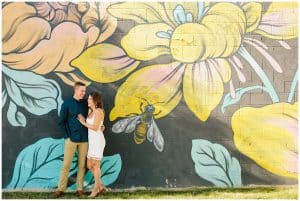 Engaged couple standing close to each other near a bright wall painted with a flower mural.
