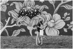 Black and white image of a couple standing in front of a flower mural. The couple are hugging each other close.