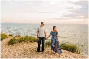 Engaged couple standing on the beach while holding hands and looking toward the water while the wind moves through their clothes.