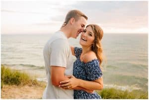 Engaged couple standing on the beach and laughing while the soon to be groom whispers into his brides ear.