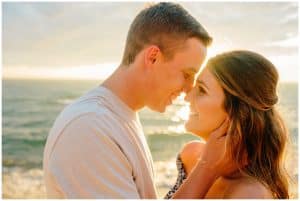 Engaged couple leaning in for a kiss with the sun peaking through between them and water sparkling in the background.