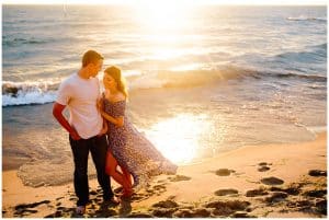 Engaged couple leaning on each other on the beach while the sun sets and makes a colorful and bright reflection on the water.