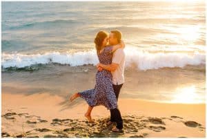Engaged couple dancing near the water on the beach at sunset. The soon to be groom is spinning his bride around while she holds onto his neck.