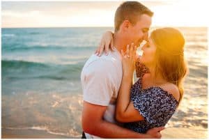 Couple leaning into each other on the beach and looking into each others eyes with sparkling water in the background.