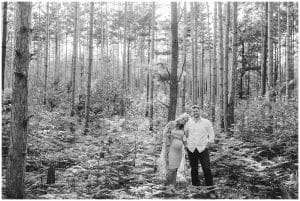 A black and white image of a couple in the forest near Riley Trails; the soon to be mother leans her head against her husbands shoulder as they both stare into the camera.