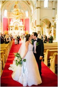 A bride and groom pause down the isle to quickly kiss one another while the audience smiles and watches.