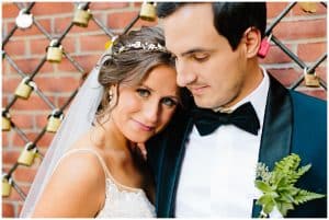 A bride and groom pose in front of a fun wall with gold accents; the bride leans her head on the grooms shoulders as he looks down on her.