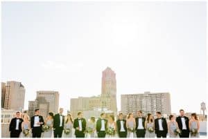 A bridal party with a city scene and tall buildings behind them as a backdrop; they are all paired up in tows and looking at eachother.