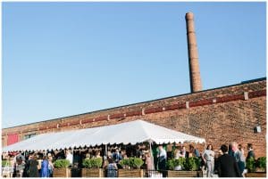 Picture of wedding guests gathering outside of a pretty brick building with a white tent; the guests are waiting for the bridal party to arrive.