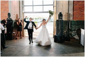 Photo of a bride and groom entering their wedding reception through an industrial style door; they have their hands raised and the bride has her flowers in the air.
