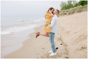 Engaged couple gets photos taken at the beach near the water. Soon to be husband picks up his bride and she wraps her hands around his neck while looking down at him.