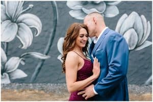 An engaged couple stands in front of a flower mural in downtown Holland. The soon to be groom leans in to kiss his future bride.