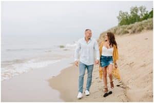 An engaged couple walk along the shoreline with water lapping up on the sand. The couple is holding hands and looking at each other.