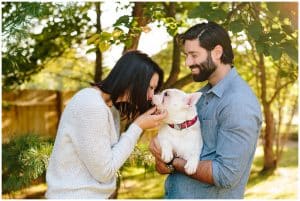 A young couple with the woman kissing their dog while the husband holds the dog. They are in a green park in early spring.