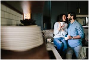 A young couple sitting on their kitchen counter for a lifestyle photo shoot. They are holding coffee and looking at each other with love in their eyes.