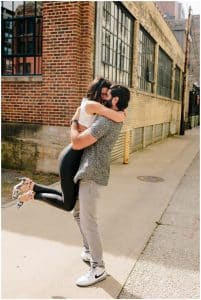 A young couple standing in front of an old building in downtown Grand Rapids while the husband picks up his wife in a big hug.