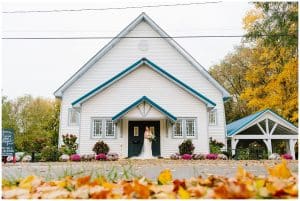 A bride stands outside a pretty white chapel with orange and red leaves sprinkling the ground.