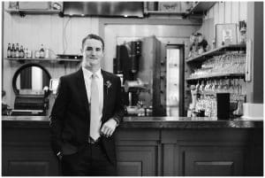 A black and white image of a groom standing in his suite in front of an old classic bar.