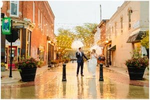 A bride and groom standing underneath an umbrella in the rain on a pretty lit street.