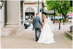 A bride and groom walk down a cobblestone street together while holding hands.