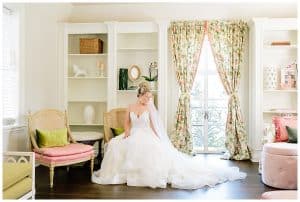 A bride poses on a vintage chair in front of a window while wearing her wedding dress.