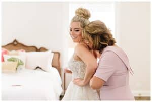 A bride smiles as her mother hugs her from behind and helps button up her wedding gown.