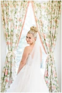 A bride turns and smiles at the camera for a picture featuring her long vail in front of a window streaming sunlight.