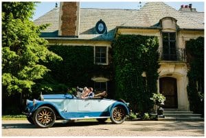 Bride and groom riding in an old blue vintage car in front of the estate they got married near.