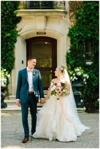 A married couple holds hands and smiles at each other for their wedding photos in front of a vintage estate building.