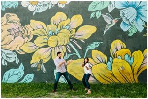 A couple makes some silly poses in front of a flower mural for their engagement photos in front of a flower mural