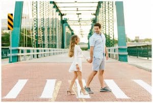 couple walks across the blue bridge in downtown Grand Rapids while holding hands and looking at each other